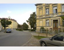 Straßenszene in einem Wohngebiet mit historischen Gebäuden, darunter ein gelbes Haus mit aufwendigen Fassadendetails und einem Holzeingangstor. Im Vordergrund geparkte Autos, Pflastersteine und ein kleiner Grünstreifen; im Hintergrund weitere mehrstöckige Wohnhäuser und Bäume.
