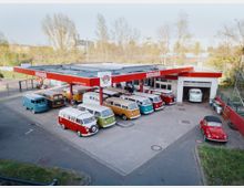 Eine ehemalige Tankstelle mit rotem Vordach, die heute als Ausstellungs- oder Werkstattfläche für VW-Bullis dient. Vor und unter dem Dach stehen verschiedene farbige Oldtimer-Bullis und ein roter VW Käfer, umgeben von einer urbanen, begrünten Umgebung.