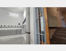 Industrieartiger Innenraum mit weiß gestrichenen Wänden und grauen Metallrohren an der Wand. Rechts befinden sich eine offene Metalltür, ein Holzrahmen, ein Heizkörper und eine Treppe, die in einen Flur führt.