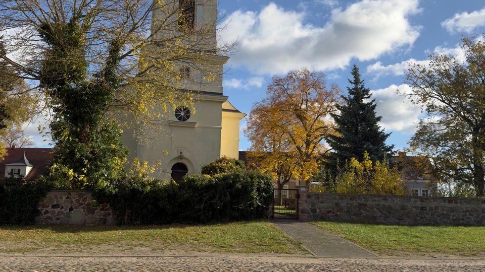 Blick auf eine helle Kirche mit quadratischem Turm und dunkler Kuppel, umgeben von Bäumen und einer niedrigen Steinmauer unter blauem Himmel mit Wolken. Im Vordergrund steht auf einem Holztisch ein Glas Kaffee, dahinter ein gepflasterter Platz.