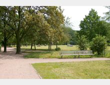 Ein Park mit breiten, sandigen Wegen, umgeben von großen Bäumen und Grünflächen. Im Vordergrund stehen zwei Holzbänke, während im Hintergrund Menschen und vereinzelte Skulpturen auf der Wiese zu sehen sind.