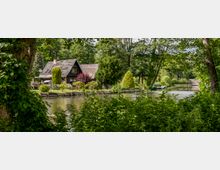 Ein idyllisches Haus mit dunkler Holzfassade und Reetdach liegt am Ufer eines ruhigen, bewachsenen Kanals. Umgeben von gepflegtem Garten und üppigen Bäumen, fügt sich das Haus harmonisch in die natürliche Umgebung ein.