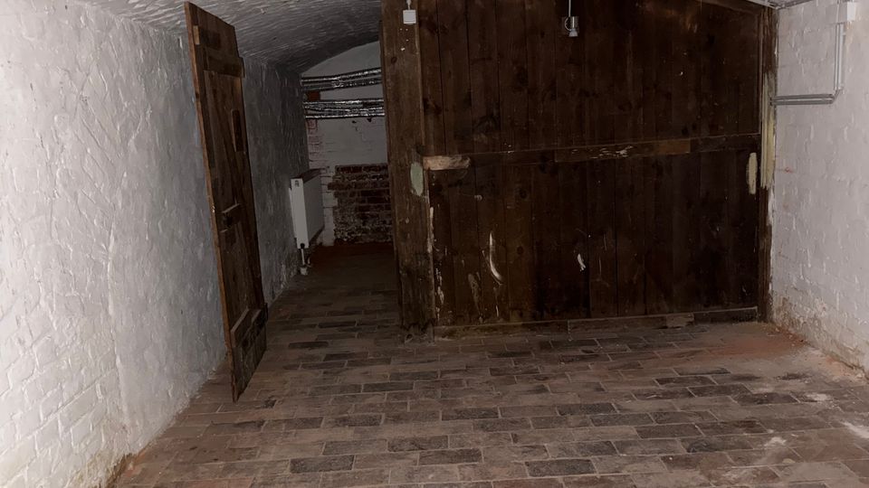 Gewölbekeller mit weiß gestrichenen Ziegelwänden und Backsteinboden, am Ende durch ein großes dunkles Holztor verschlossen. Links steht eine geöffnete Holztür zu einem schmalen Gang mit freiliegenden Leitungen und einem Heizkörper.
