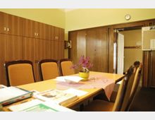 Ein Besprechungsraum mit einem rechteckigen Holztisch, umgeben von gepolsterten Stühlen. An der Wand befinden sich hölzerne Einbauschränke und eine Tür führt in einen angrenzenden Flur.