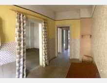 Ein heller Innenraum mit gelb gestrichenen Wänden, teils bedeckt mit gemusterten Tapeten und Vorhängen mit buntem Design. Zwei Türöffnungen führen in angrenzende leere Räume, während ein kleiner Holzstuhl und ein schmaler Tisch im Vordergrund sichtbar sind.