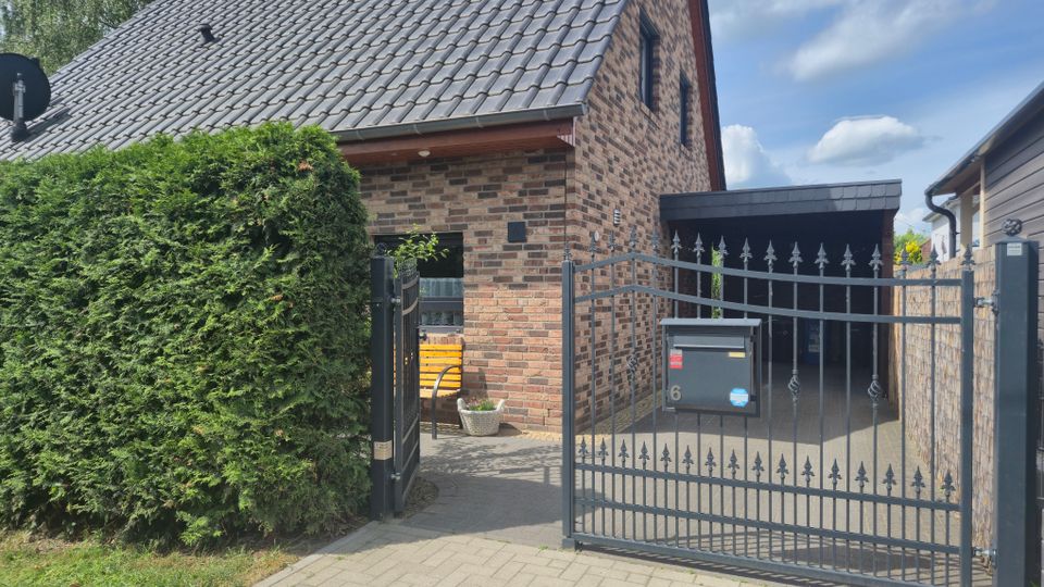 Einfamilienhaus mit Ziegelfassade und steilem, grauem Ziegeldach, davor ein schmiedeeisernes Tor mit Briefkasten und Zufahrt zu einem überdachten Carport. Links steht eine hohe, dicht geschnittene Hecke, und der Gehweg führt zum Eingang unter dem Dachüberstand.