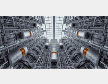 Innenansicht eines modernen Gebäudes mit mehreren gläsernen Aufzugskabinen, die in einer symmetrischen Anordnung zwischen Stahlgerüsten verlaufen. Oben ist ein Dach aus gläsernen Paneelen zu sehen, das Tageslicht in den Raum einlässt.