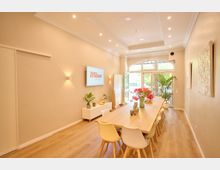 Heller Besprechungsraum mit einem langen Holztisch und bunten Stühlen, umgeben von beige gestrichenen Wänden. Im Hintergrund ein großer, bogenförmiger Glaseingang mit Blick ins Grüne, Pflanzen und Dekorationen ergänzen das moderne Interieur.