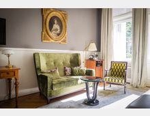 Wohnzimmer mit klassischer Einrichtung: Ein grünes Sofa mit Zierkissen, ein gelb-gemusterter Sessel, ein kleiner runder Glastisch und ein Holz-Beistelltisch mit Lampe. Im Hintergrund ein Porträt in einem goldenen Rahmen an der grau gestrichenen Wand und ein großes Fenster mit weißen Vorhängen, durch das Tageslicht einfällt.