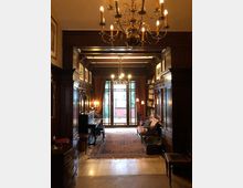 Ein gemütlicher Salon mit dunklen Holzvertäfelungen, einem Kronleuchter an der Decke, einem Klavier und Bücherregalen. Große Fenster mit Vorhängen im Hintergrund bieten Blick auf einen Garten, und der Raum ist mit Teppichen und eleganten Möbeln eingerichtet.