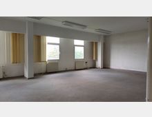 Leerer Raum mit weißen Wänden und großen Fenstern mit vertikalen, gelblichen Lamellen. Der Raum hat einen grauen Teppichboden, sichtbare Heizkörper unter den Fenstern und Deckenleuchten im rechteckigen Format.