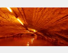 Backstein-Tunnelgewölbe mit warm orangefarbenem Licht beleuchtet, unter dem Wasser fließt; entlang der Decke verlaufen Rohre und Kabel.