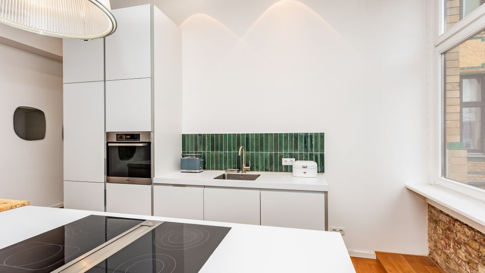 Moderne, minimalistische Küche mit weißen, griffarmen Schränken, eingebautem Backofen und einer Arbeitszeile mit Spüle vor einer grünen Fliesenrückwand. Im Vordergrund steht eine Kochinsel mit Induktionskochfeld; rechts sorgt ein großes Fenster mit gemauerter Brüstung und Holzstufen für Tageslicht.