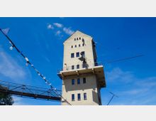 Ein mehrstöckiger, beige-grauer Turm mit kleinen Fenstern und einer umlaufenden Plattform in der oberen Etage, vor einem blauen Himmel. Links ist eine Fußgängerbrücke erkennbar, daneben hängen Wimpelketten mit Lichtern in der Luft.