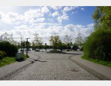 Gepflasterter Platz mit einem zentralen Rundbeet, umgeben von Bäumen, in direkter Nähe zu einem Fluss mit einer Steinbrücke im Hintergrund. Rechts und links säumen Gehwege sowie grüne Büsche und Rasenflächen die Szene, während der Himmel mit einigen Wolken bedeckt ist.