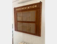 Holzgerahmter Schaukasten an einer weißen Innenwand, oben mit dem Schriftzug „DOKUMENTATION“. Hinter Glasscheiben sind leere Korkflächen in mehreren Feldern zu sehen, mit sichtbaren Befestigungsschrauben.