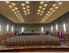 Ein mittelgroßer Saal mit Holzklappstühlen in mehreren Sitzreihen, einer Bühne im Vordergrund und einer Rückwand mit vertikal gerilltem Design. Die Decke ist mit einer regelmäßigen Anordnung von kleinen gelben Leuchten verziert; an den Wänden befinden sich mehrere Wandlampen. Im Hintergrund sind Personen zu sehen, die sich unterhalten.