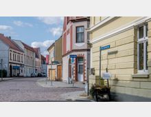 Gepflasterte Straßenszene in einer Altstadt mit zweigeschossigen Häusern, teils mit verzierten Fassaden in Beige- und Rottönen. Auf der rechten Seite ein gelbes Gebäude mit Straßenschildern wie „Alte Kirchstraße“ und „Königsstraße“ sowie ein Wegweiser zu einem Sonnengarten.