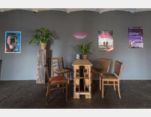 Ein minimalistischer Raum mit einer grauen Wand, an der Filmplakate und ein Motivationsspruch angebracht sind. In der Mitte stehen ein schmaler Tisch aus Holzpaletten, vier verschiedene Holzstühle und Topfpflanzen, die den Raum dekorieren.