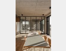 Moderner Besprechungsraum mit einer großen rechteckigen Tischfläche und Holzstühlen auf einem hellen Holzboden. Der Raum hat Glaswände, Sichtbetondecken und großen Fensterfronten, die Tageslicht hereinlassen und einen Blick auf die umliegende Landschaft ermöglichen.