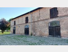 Backsteingebäude mit großflächigen Holztoren und Fenstern mit Sprossenglas, umgeben von einem gepflasterten Hof und vereinzeltem Grün. Links im Bild stehen Bäume, die teilweise Schatten spenden.