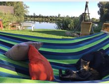 Eine Hängematte in Blau- und Grüntönen mit Kissen und einer Katze darauf, auf einer Wiese mit Blick auf einen ruhigen See im Hintergrund. Im Umfeld sind ein weißer Gartenstuhl, ein Holzgestell sowie Schilfpflanzen und Bäume am Ufer zu sehen.