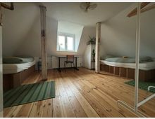 Dachzimmer mit Holzboden, zwei einfachen Betten auf niedrigen Boxen, einem Schreibtisch und Stuhl unter einem Fenster. Zwei sichtbare Holzstützbalken und minimalistische Einrichtung mit Pflanzen sowie einem Kleiderständer.