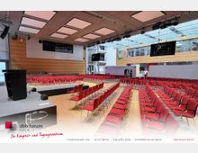 Moderner Konferenzraum mit einem erhöhten Podium, roten Polsterstühlen in Reihen angeordnet, und einer großen Glasdecke, die viel Licht einlässt. Hinten sind eine Holzwand und eine verglaste Galerie sichtbar.