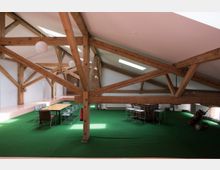 Großzügiger Dachraum mit sichtbaren Holzbalken und grünem Teppichboden. In der Mitte steht ein langer Tisch mit Stühlen, daneben befinden sich weitere Tische und ein Golf-Caddy; Tageslicht fällt durch dachhohe Fenster und Oberlichter ein.