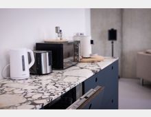 Eine moderne Küchenzeile mit einer Arbeitsplatte aus weiß-schwarz gemustertem Marmor, darauf befinden sich ein Wasserkocher, ein Toaster und eine Mikrowelle. Im Hintergrund ist eine graue Betonwand sichtbar, daneben ein Teilbereich eines Sofas.
