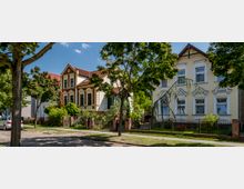 Straßenszene mit zwei historischen Wohnhäusern, die sich durch verzierte Fassaden mit weißen und gelben Elementen auszeichnen. Im Vordergrund sind Bäume, ein Gehweg und ein gepflasterter Straßenabschnitt sichtbar, während ein schmiedeeisener Zaun die Grundstücke umgibt.