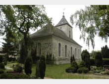 Steinkirche mit Satteldach und kleinem Glockenturm; das Gebäude hat bogenförmige Fenster und steht inmitten eines gepflegten Friedhofs mit Hecken, Bäumen und Blumen. Im Hintergrund sind weitere Gräber und Bäume sichtbar.