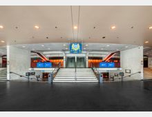 Weitläufiger, moderner Eingangsbereich eines Gebäudes mit Treppen in der Mitte, die zu einer großen Doppeltür führen. Betonwände, ein heller Deckenbereich mit eingebauten Leuchten, ein blauer Orientierungsschilder und digitale Bildschirme, die an den Seiten befestigt sind, prägen die Architektur.