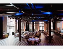 Großer, industrieller Veranstaltungsraum mit hohen Decken, sichtbaren Stahlträgern und großen Fabrikfenstern. Der Raum ist für eine Feier dekoriert, mit runden und rechteckigen Tischen, weißen Tischdecken und Lichterketten an den Balken.