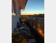 Blick auf einen Balkon mit Sitzkissen, kleinen Pflanzen und einer Kerze, der sich an der Seite eines Mehrfamilienhauses befindet. Im Hintergrund ist eine Stadtsilhouette unter einem Himmel bei Sonnenuntergang zu sehen.