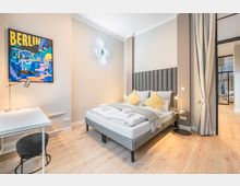 Modernes Schlafzimmer mit Doppelbett, hohem gepolstertem Kopfteil, Nachttischlampen und hellem Holzboden. Links steht ein weißer Schreibtisch unter einem „Berlin“-Poster, rechts trennt eine Glaswand mit Vorhang den Raum zu einem Flur oder Nebenbereich.