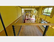 Innenraum eines gelben Veranstaltungsraums mit hohen Decken, großen Fenstern und dekorativen Kronleuchtern. Der Raum verfügt über eine Holzbrüstung auf einer Galerie und einen Bereich mit zusammengestellten Tischen und roten Stühlen auf einem Parkettboden.