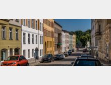 Pflastersteinstraße in einer Wohngegend mit farbigen mehrstöckigen Gebäuden, teils renoviert, teils historisch, auf der linken Seite. Auf beiden Straßenseiten parken Autos, im Hintergrund sind Bäume eines Parks oder Waldes zu sehen.