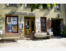Fassade eines kleinen Kinos mit dem Namen "Lichtblick Kino" in einem grünen Gebäudeeingang. Die Außenwand ist mit Plakaten, Graffiti und angeklebten Zetteln bedeckt, während zwei Holzbänke und ein Kopfsteinpflaster den Eingangsbereich säumen.