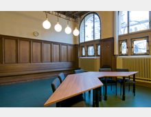 Ein historischer Besprechungsraum mit holzverkleideten Wänden, einer Bank entlang der Wand und großen Bogenfenstern mit Sprossen. Ein moderner, dreieckiger Besprechungstisch mit Stühlen steht auf einem grünen Boden, und eine Hängeleuchte mit vier großen, runden Lampen ist an der Decke angebracht.