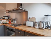 Moderne Küche mit L-förmiger Arbeitsplatte aus Holz, Herd mit eingebautem Backofen, Abzugshaube und Edelstahlrückwand. Auf der Arbeitsfläche stehen diverse Küchengeräte, darunter ein Toaster, Wasserkocher, Mixer und Gewürzgläser, sowie Kochutensilien in einem Halter.