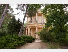 Historisches Gebäude mit runder Architektur und Säulengang, umgeben von Bäumen, Sträuchern und einem schmalen Sandweg im Vordergrund. Das Gebäude hat cremefarbene Fassade und hohe Fenster.