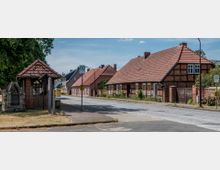 Kleiner Dorfplatz mit einer Bushaltestelle, einer überdachten Backsteinkonstruktion im Vordergrund und Fachwerkhäusern mit roten Ziegeldächern entlang einer gepflasterten Straße. Im Hintergrund sind weitere Wohnhäuser und Bäume zu sehen.