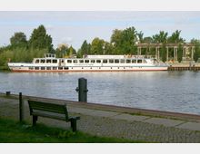 Ein weißes Ausflugsschiff mit der Aufschrift "Charlottenhof" fährt auf einem Fluss, umgeben von grünen Bäumen und historischen Säulenstrukturen im Hintergrund. Im Vordergrund ist eine Uferpromenade mit einer Bank und gepflasterten Wegen zu sehen.