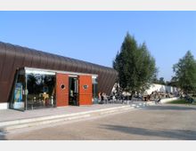 Ein modernes Gebäude mit einer rundlichen Metallfassade und großen Glasflächen, dessen Eingangsbereich aus zwei offenen Holztüren besteht. Davor befinden sich eine Terrasse mit Sitzgelegenheiten, sowie Bäume und ein Gehweg mit parkenden Fahrrädern im Hintergrund.