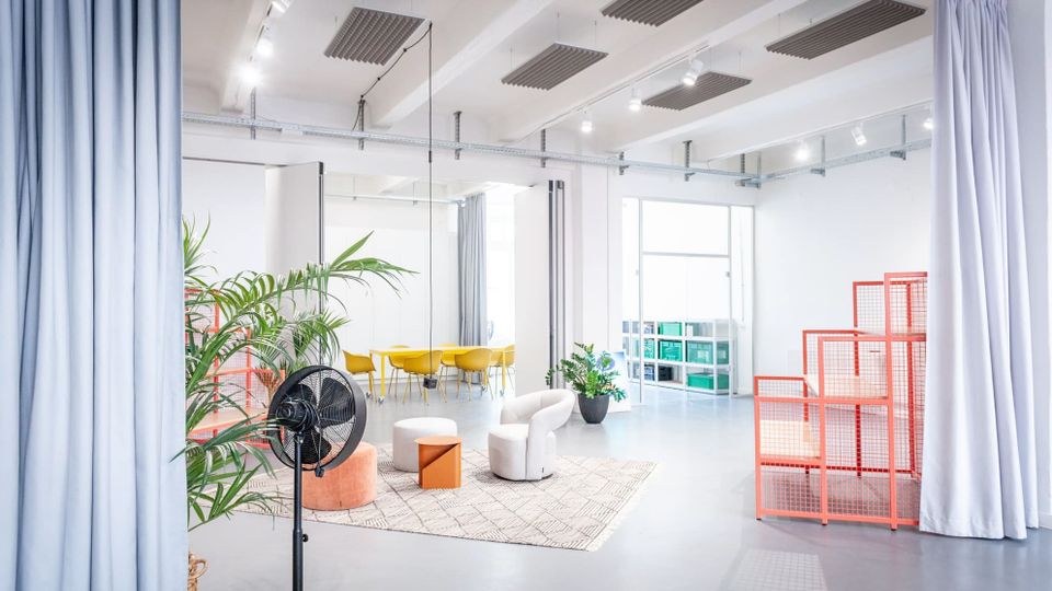 Heller, offener Innenraum eines modernen Studios oder Coworking-Spaces mit hohen Decken, Akustikpaneelen und Vorhängen als Raumteiler. In der Mitte stehen Sitzmöbel auf einem Teppich, dahinter ein gelber Tischbereich, Pflanzen, ein Regal und rote Gitterpodeste neben großen Fenstern.