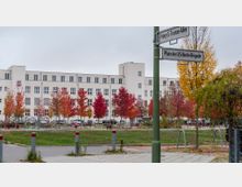 Straßenkreuzung mit einem weißen, mehrstöckigen Gebäude im Hintergrund, umgeben von bunten Bäumen in Herbstfarben. Im Vordergrund befinden sich eine kleine Grünfläche, Spielgeräte und Straßenschilder mit den Namen „Harry-S.-Truman-Allee“ und „Platz der US-Berlin-Brigade“.
