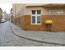 Pflastersteinstraße in einem Wohngebiet mit einem Gebäude mit braunen Fliesenfassaden. An der Ecke des Gebäudes befinden sich ein gelber Briefkasten und ein Schild mit der Aufschrift „Krankentransport Haeberer“.