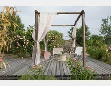 Holzterrasse mit einem einfachen Pergolagerüst aus Holz, an dem helle Stoffbahnen befestigt sind. Darunter stehen Gartenmöbel, darunter ein runder Tisch und ein Schaukelstuhl, umgeben von blühenden Rosenbüschen und grüner Vegetation in einer natürlichen Gartenlandschaft.