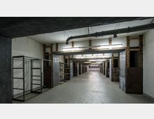 Langer Raum mit industriellem Charakter, gekennzeichnet durch freiliegende Holzbalken, metallene Regale und weiße Wände. Die Decke zeigt sichtbare Rohrleitungen, und der Raum wird durch Leuchtstoffröhren beleuchtet.